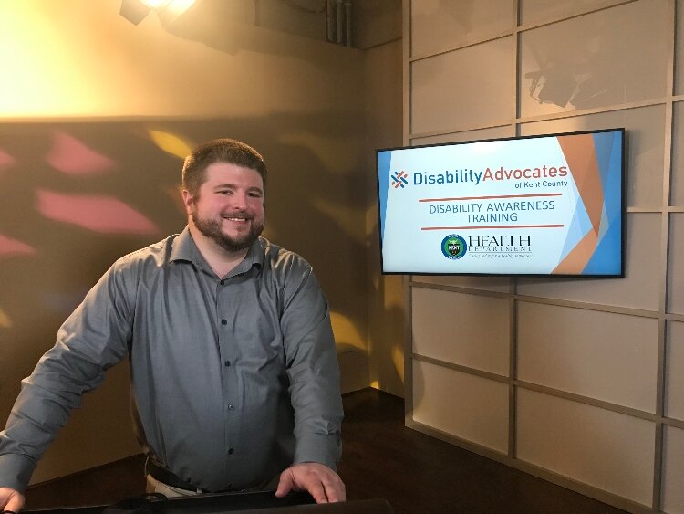 Jon Cauchi is a DEIA speaker for speaker for Disability Advocates of Kent County. In the photo, Jon Cauchi, who has a beard and is wearing a gray shirt, is standing in front of monitor advertising disability awareness training. 