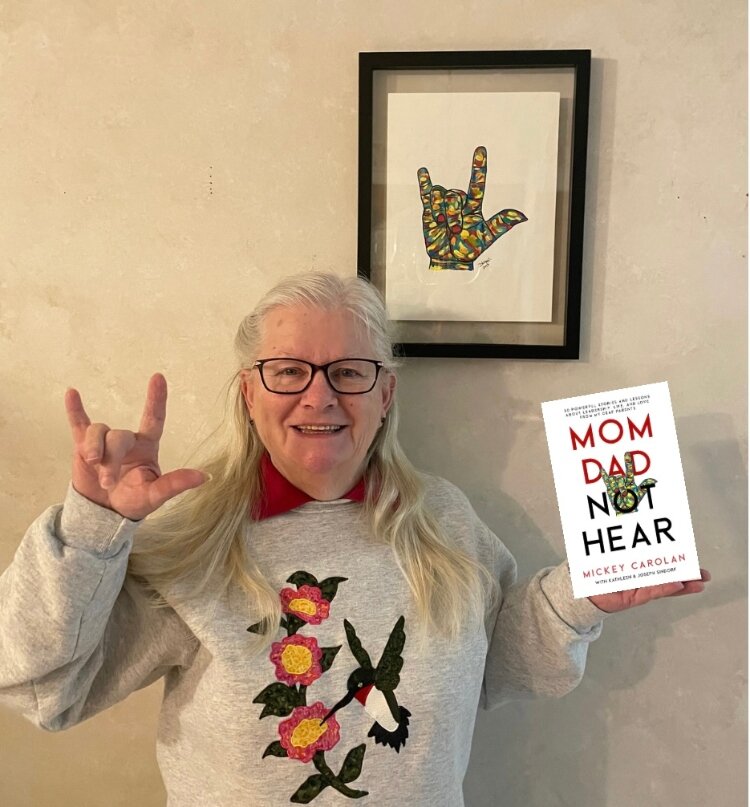 Bonnie Carolan holding her son's book about her and saying "I love you" in ASL.